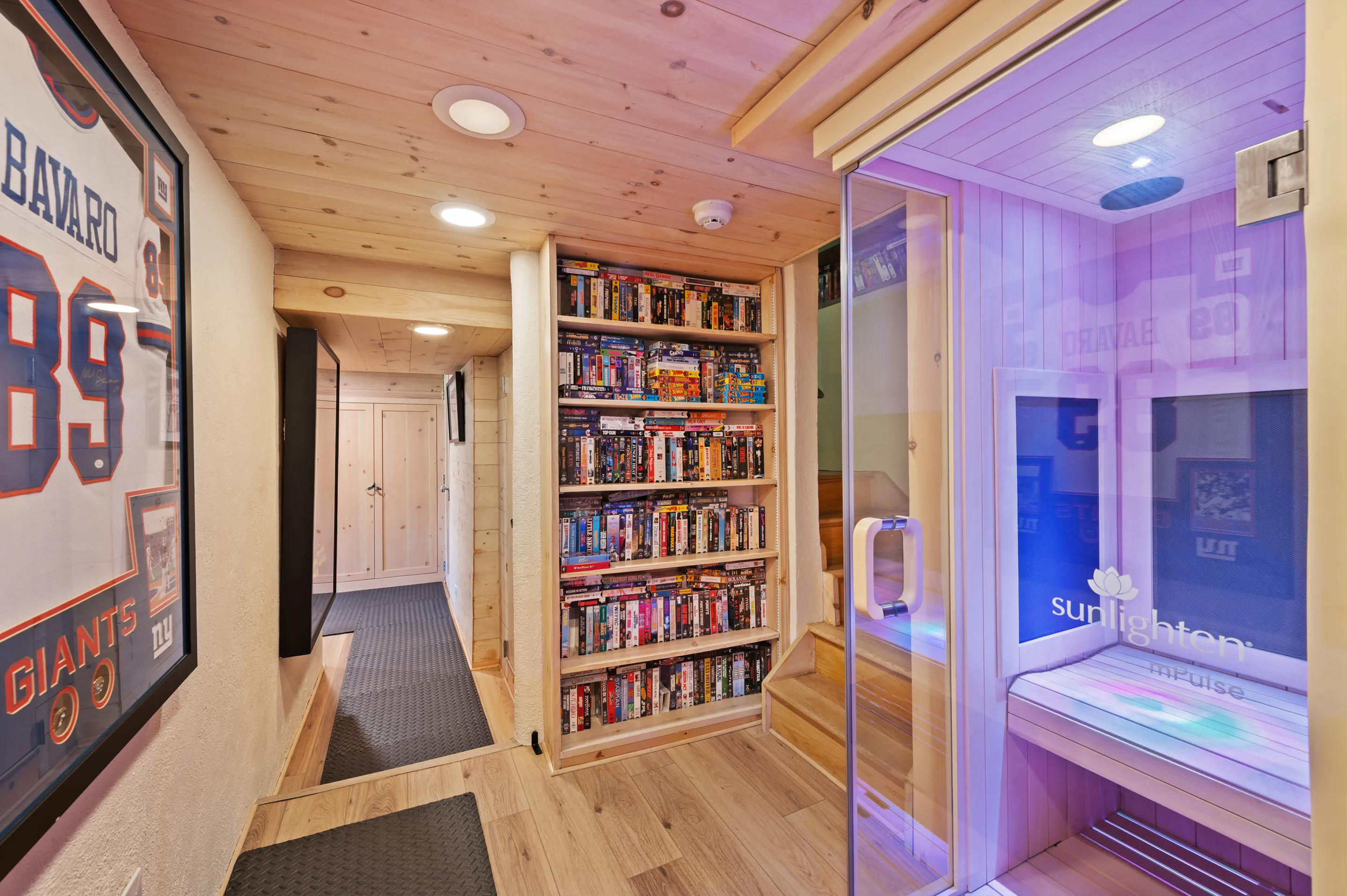 A room with light wood paneled walls and ceiling, a full bookcase of VHS tapes, a glass-encased sauna with purple lighting, and a framed sports jersey.