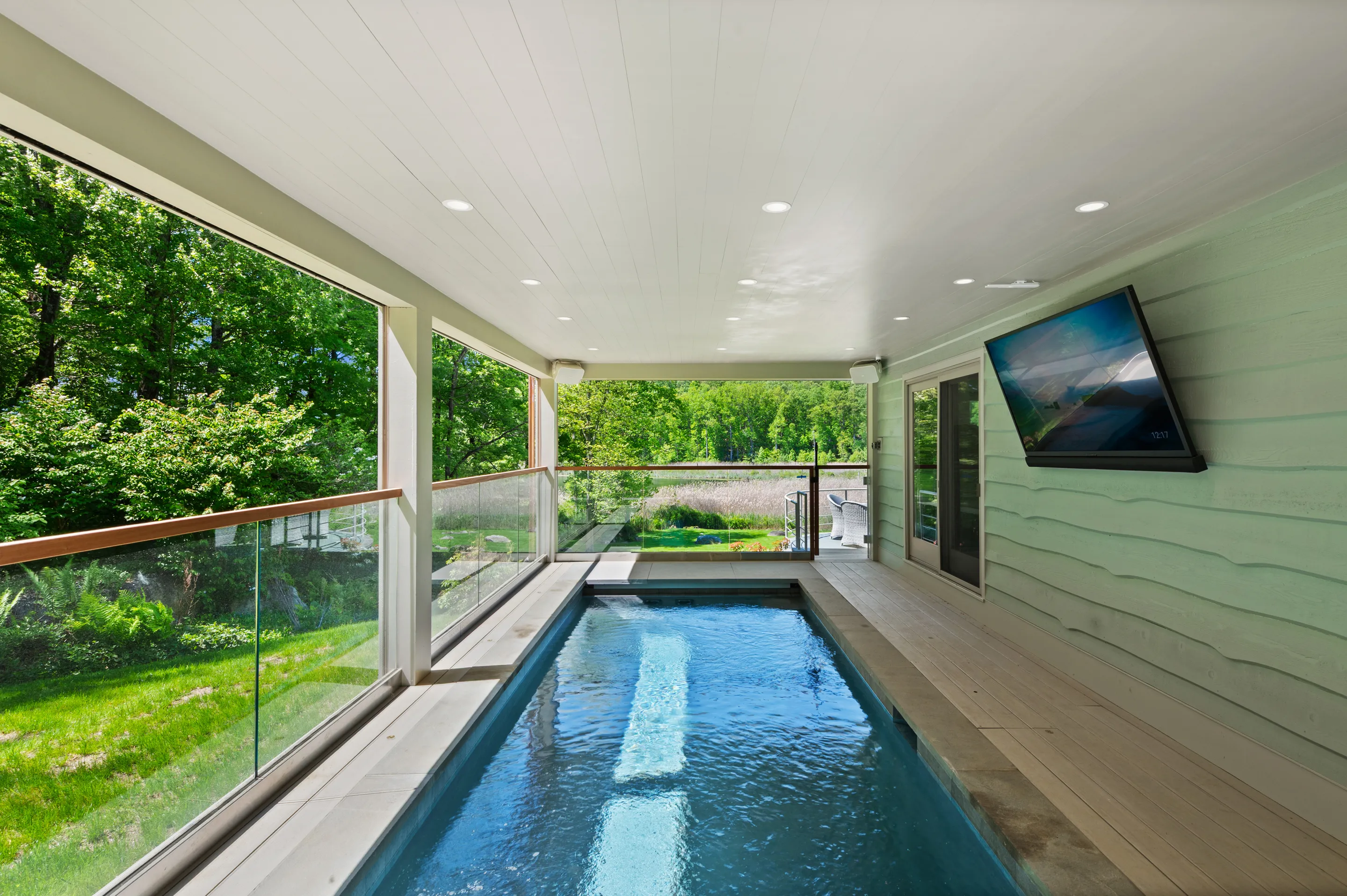 Indoor-outdoor pool on a covered patio with a TV mounted on a light green wall and lush greenery visible through glass railings.