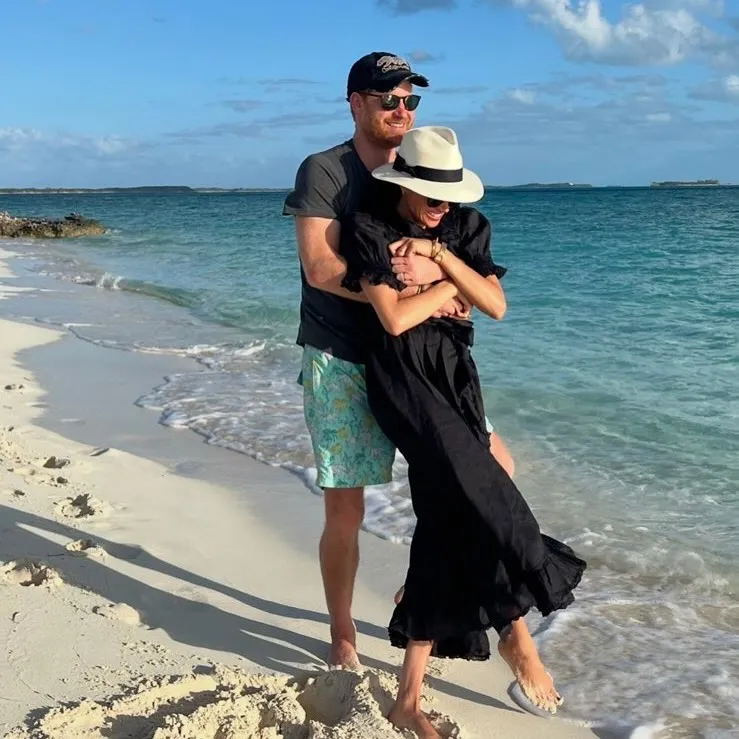 Meghan Markle and Prince Harry embracing on a sandy beach with blue water.