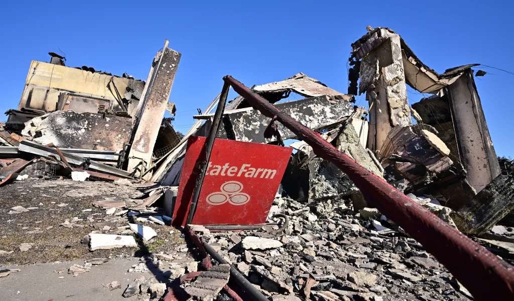A red State Farm sign stands amid the rubble of a building destroyed by the Palisades Fire.
