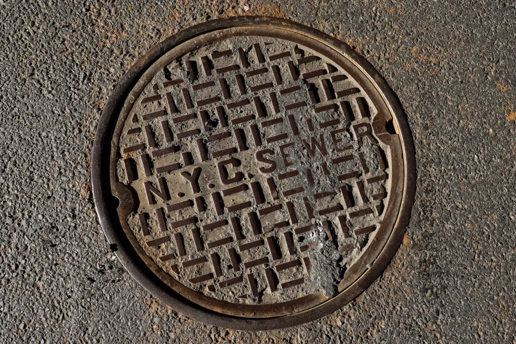 A circular metal manhole cover embedded in asphalt, inscribed with "N.Y.C. SEWER" in block letters.
