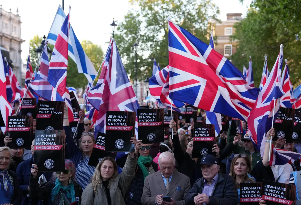 People at a rally against antisemitism holding signs that read "Jew-hate is now a national emergency" and Union Jack flags.