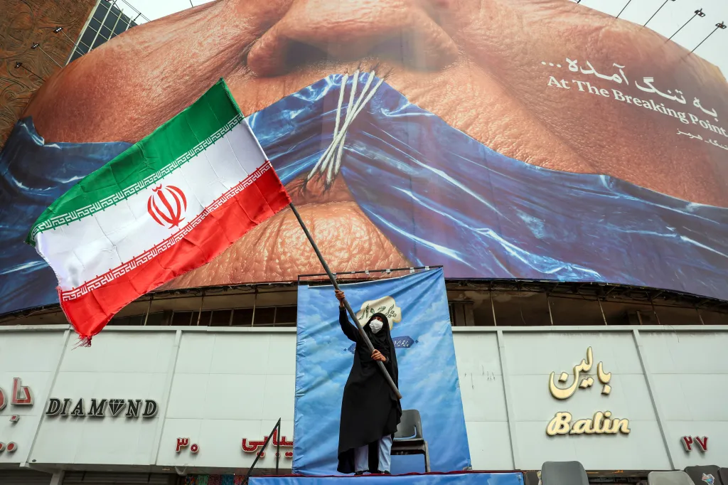 A woman in a black chador and white mask holds an Iranian flag in front of a billboard with a giant image of a person's face, partially obscured by blue fabric, and text in English and Farsi that reads 