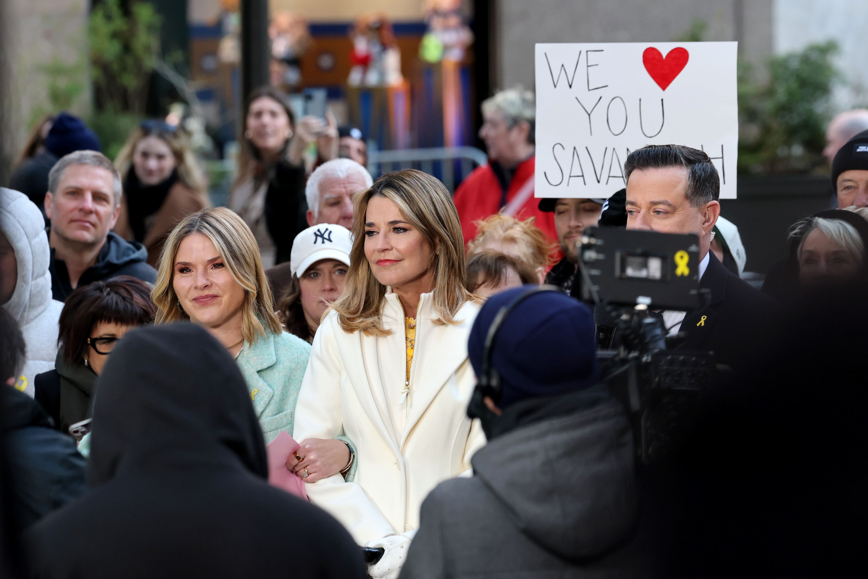 Savannah Guthrie and Jenna Bush Hager among a crowd on the "Today" show plaza, with a sign in the background reading "WE ❤️ YOU SAVANNAH".