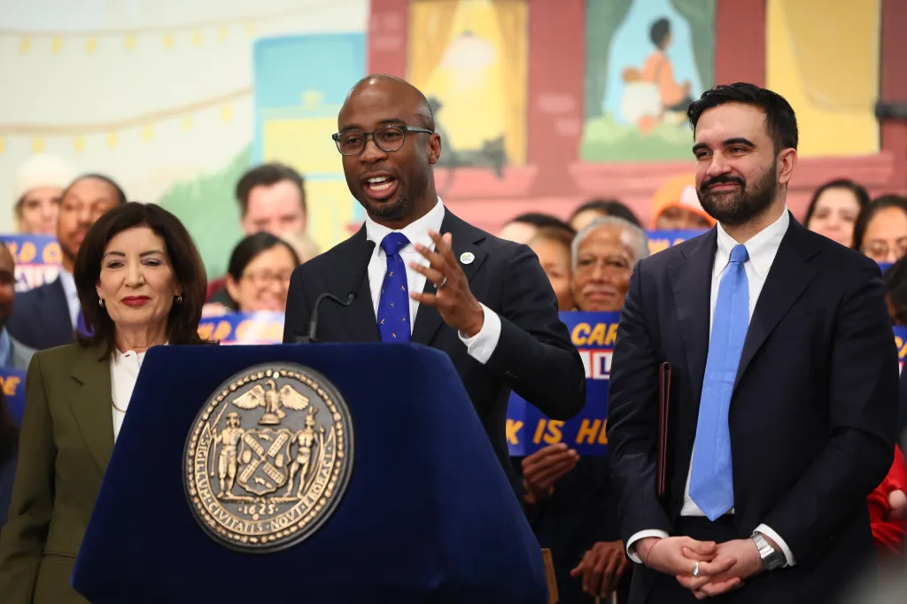 Kamar H. Samuels speaking at a podium next to New York Governor Kathy Hochul and Mayor Zohran Mamdani.