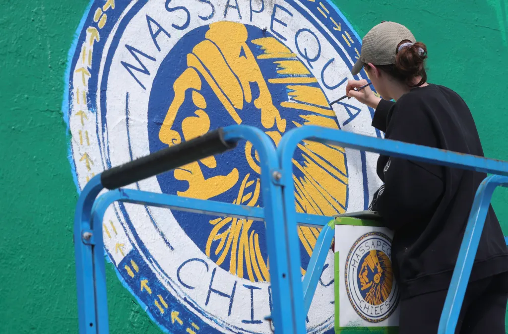 A student from Massapequa High School painting a mural with the school's logo, which features a Native American, on a green wall.