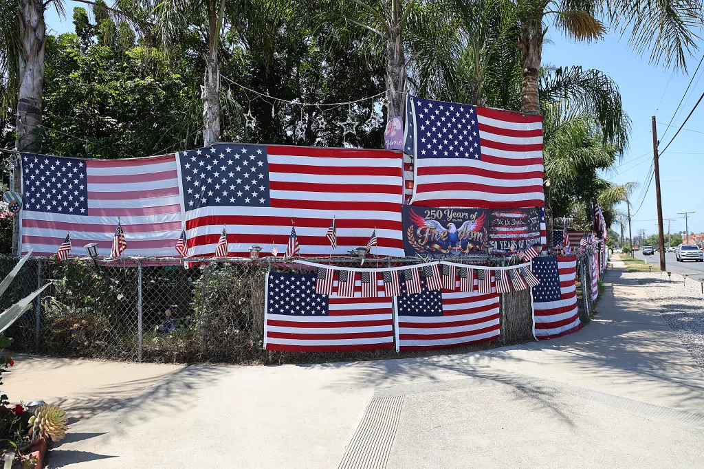 Numerous American flags and banners celebrating "250 Years of Freedom" adorn a chain-link fence alongside a sidewalk and road.