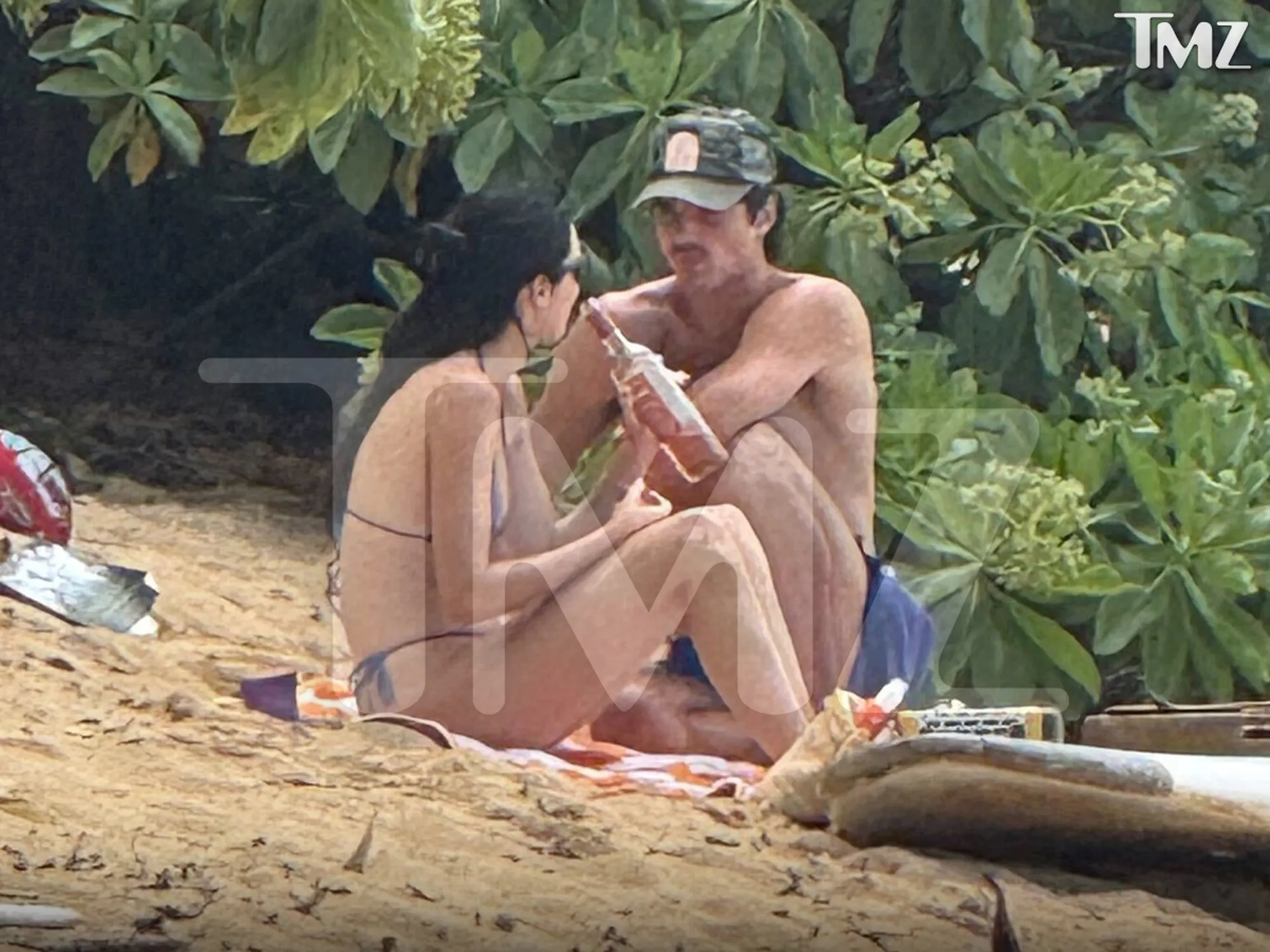 Kendall Jenner and Jacob Elordi sitting on a beach towel on a sandy beach, surrounded by green foliage, with Jacob holding a bottle.