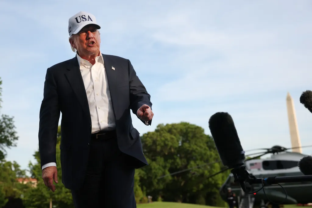 President Trump, wearing a "USA" cap, speaks to the media with the Washington Monument and Marine One in the background.