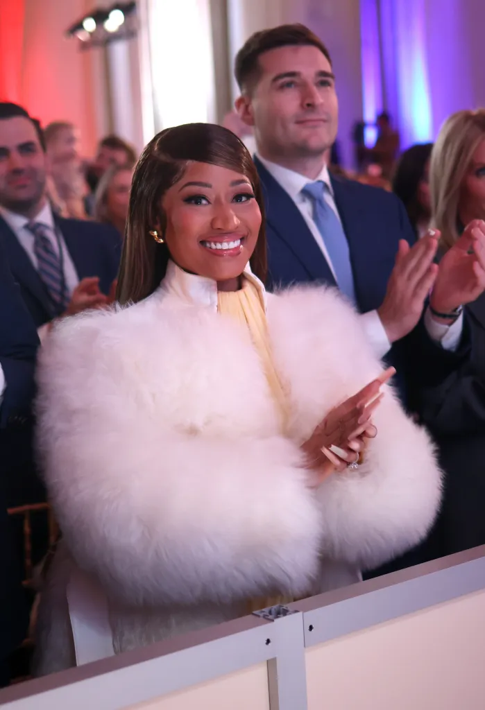 Nicki Minaj smiling and clapping while wearing a white fur coat and yellow dress.