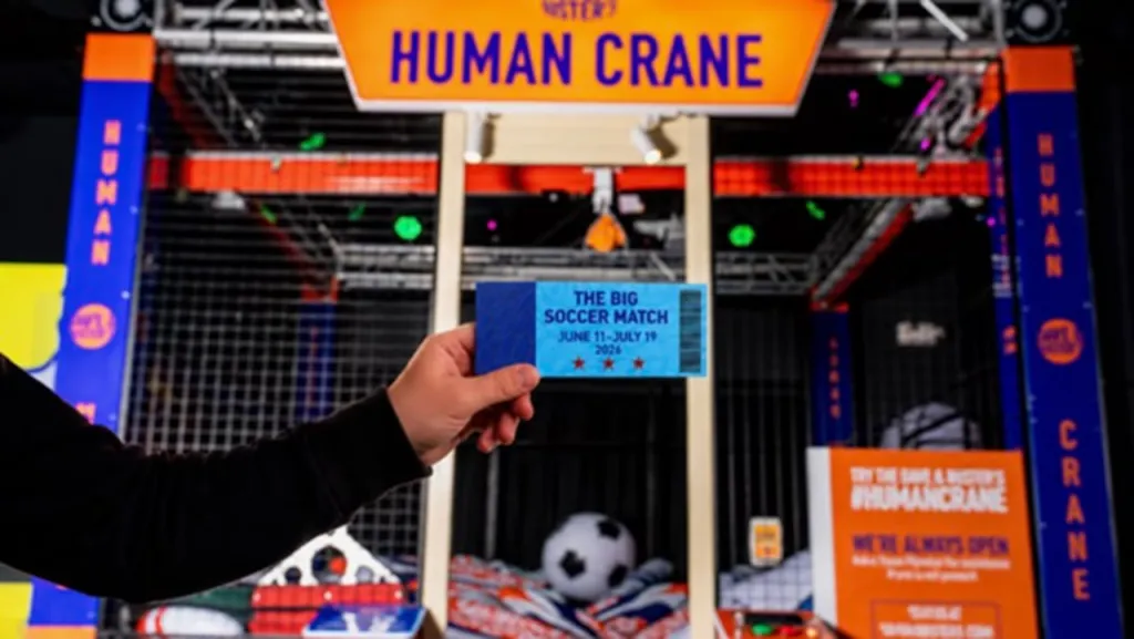 A hand holding a ticket to "The Big Soccer Match" in front of a "Human Crane" arcade game.