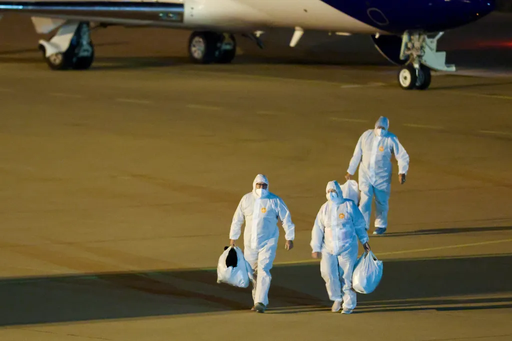 Three people in white hazmat suits and face masks carry white bags on an airport tarmac at night, with an airplane visible in the background.