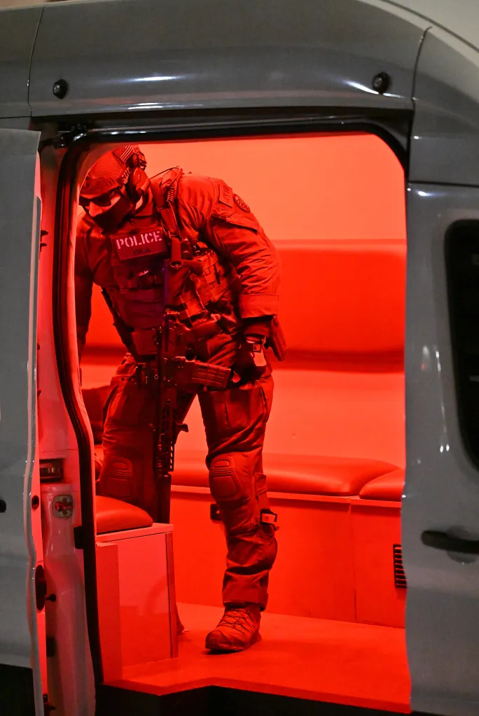 A tactical police officer wearing a uniform and carrying a rifle exits a vehicle, illuminated by red light.
