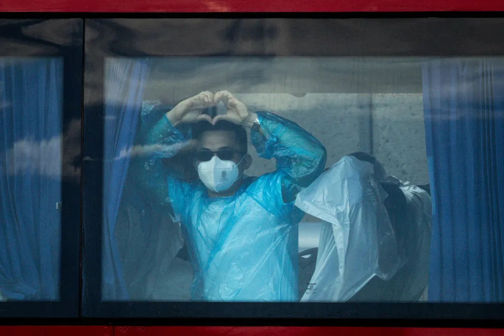 A person in a face mask, sunglasses, and a plastic medical gown makes a heart shape with their hands while looking out of a window.