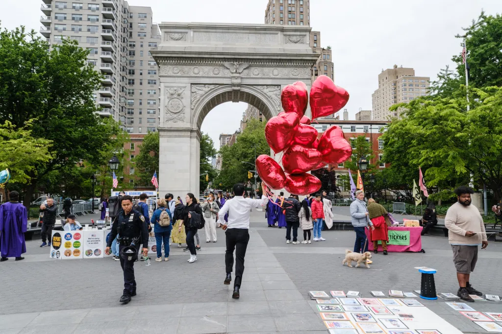 A man carries a large bunch of red heart balloons while walking past the Washington Square Arch, a New York City Police Department officer, and a political booth with