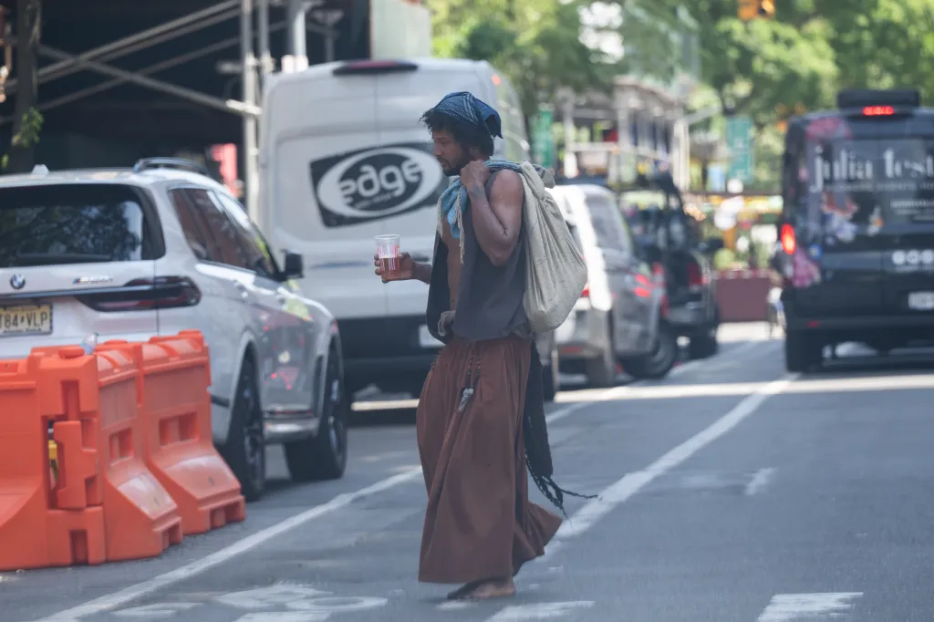 A man, barefoot and dressed in brown pants and a dark vest with a light-colored bag over his shoulder, walks down a street holding a plastic cup.