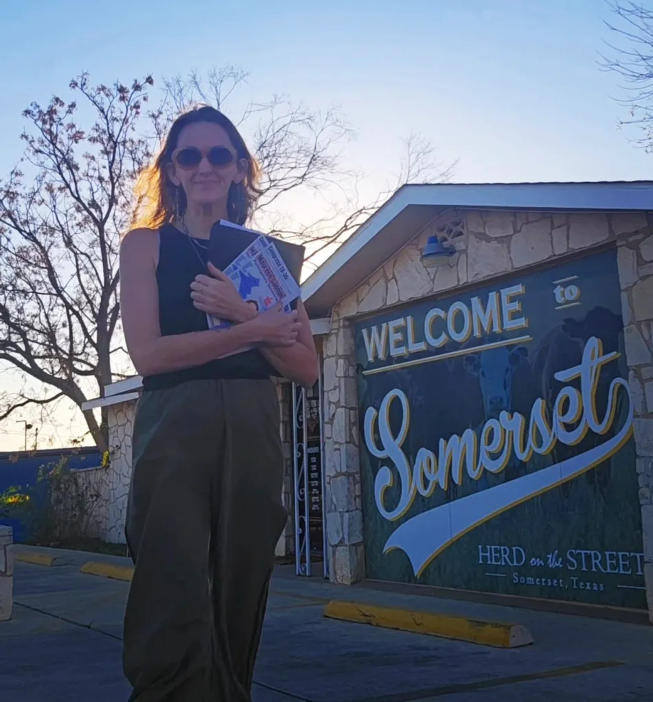 A woman holding campaign flyers for Maureen Galindo stands in front of a "Welcome to Somerset, Texas" sign.