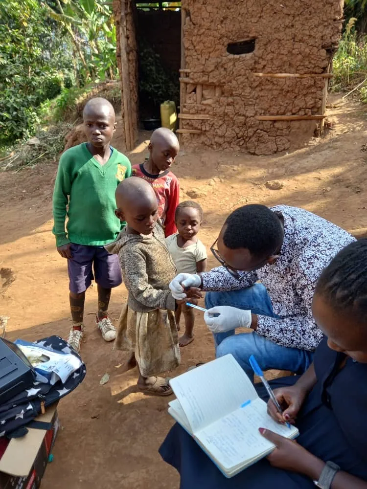 Two health workers test children while writing notes in a notebook on a dirt road.