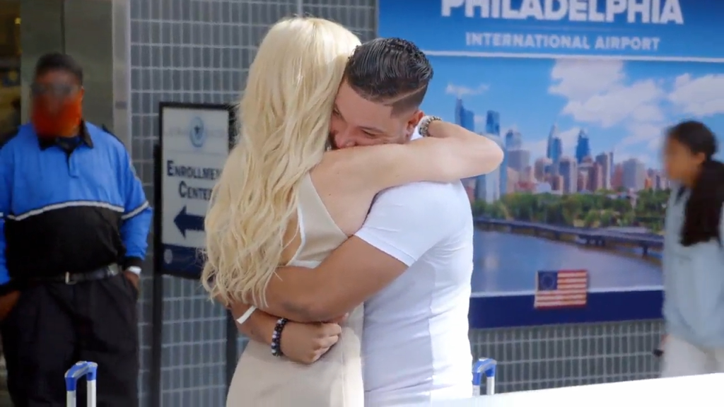 A blonde woman and a man in a white shirt embrace at an airport, with a "Philadelphia International Airport" sign in the background.