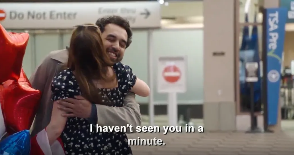A smiling man embraces a woman holding red and blue balloons at an airport, with the text 