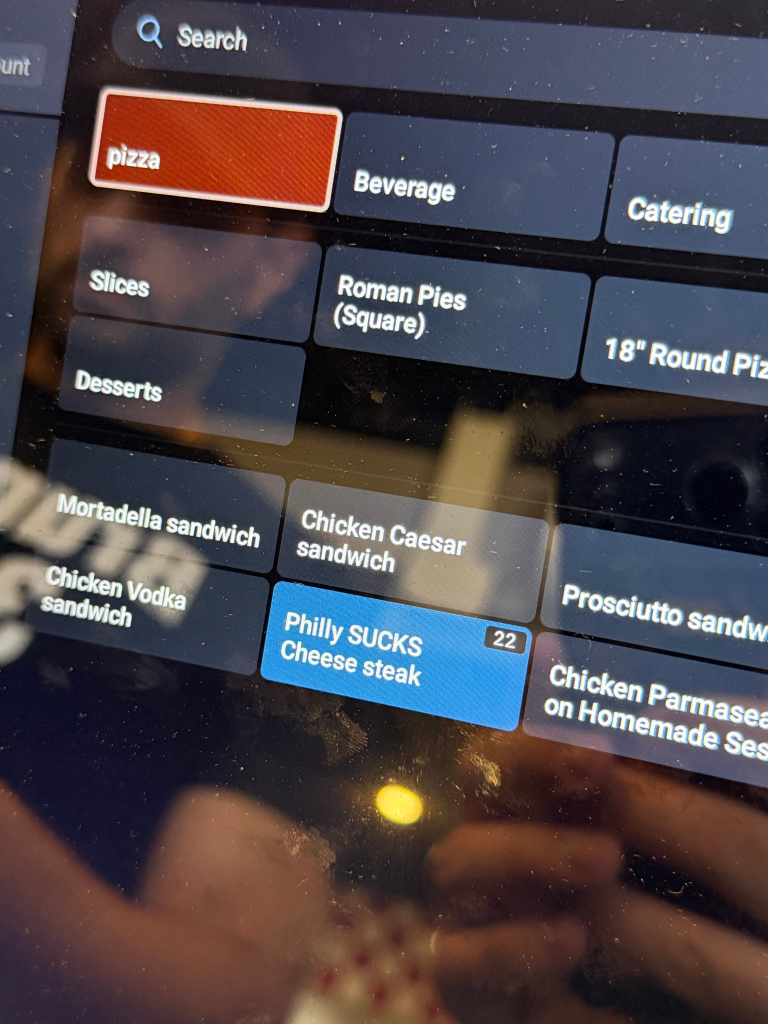 A digital menu on a screen displaying menu categories such as "pizza", "Beverage", "Catering", "Slices", "Roman Pies (Square)", "Desserts", "18" Round Pizzas", "Mortadella sandwich", "Chicken Caesar sandwich", "Prosciutto sandwich", "Chicken Vodka sandwich", and "Philly SUCKS Cheese steak".