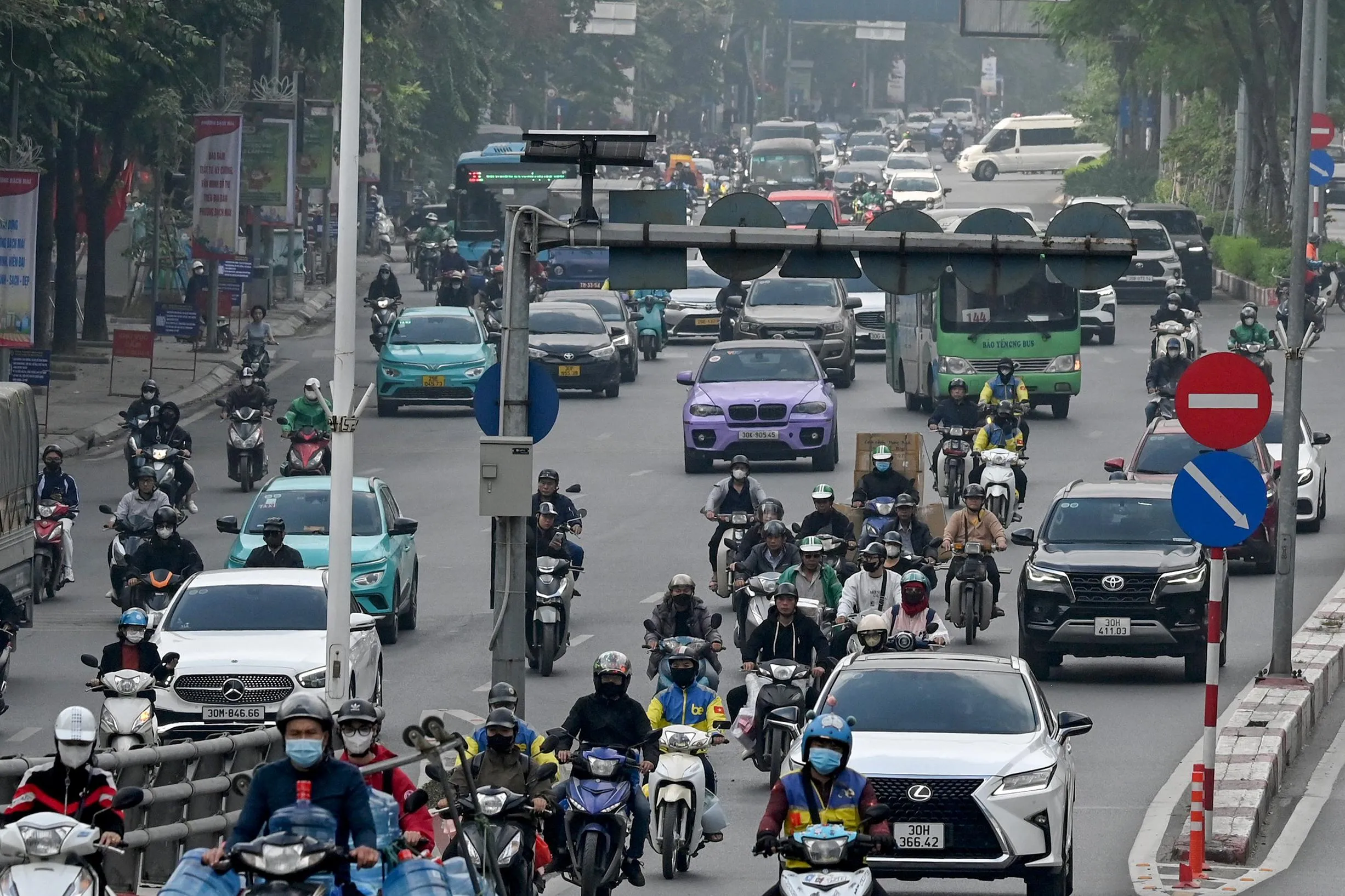 Dense urban traffic of motorbikes, cars, taxis, and buses fills a hazy multilane street, with riders packed closely together in mixed traffic.