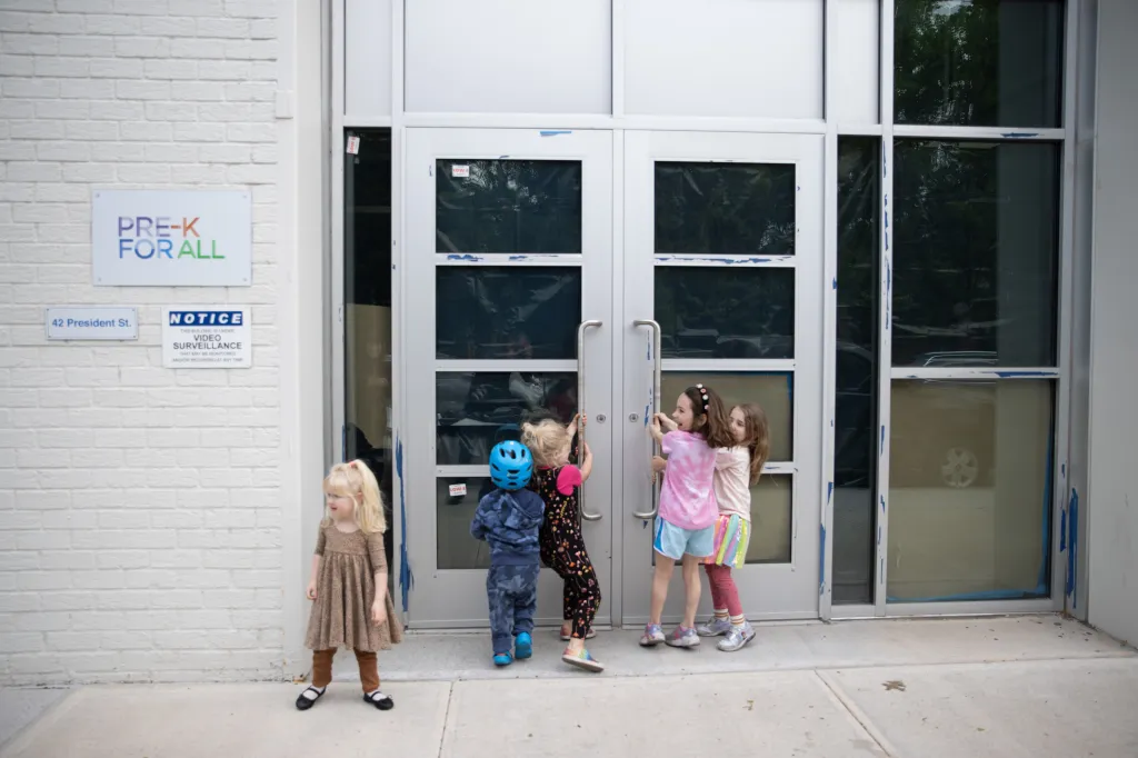 Five young children outside a building with a "Pre-K for All" sign and a "Video Surveillance" notice.