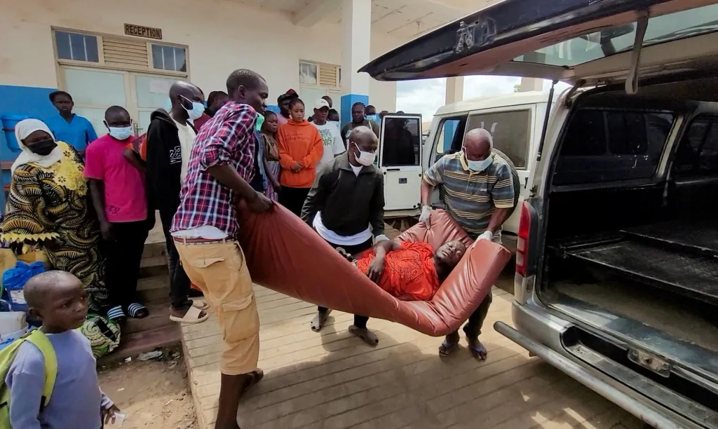 People carry a patient on a stretcher toward an ambulance as others look on outside a building with a "Reception" sign.
