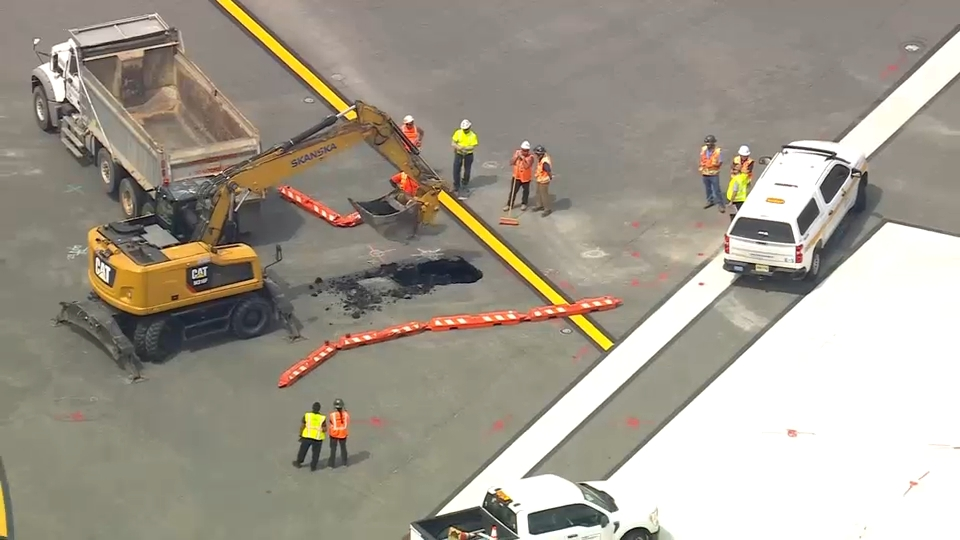 An aerial view of a sinkhole on an airport runway with an excavator, dump truck, utility vehicles, and several workers in hard hats and safety vests.