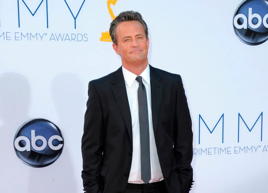 Matthew Perry in a black suit with a gray tie, smiling at the 64th Primetime Emmy Awards.