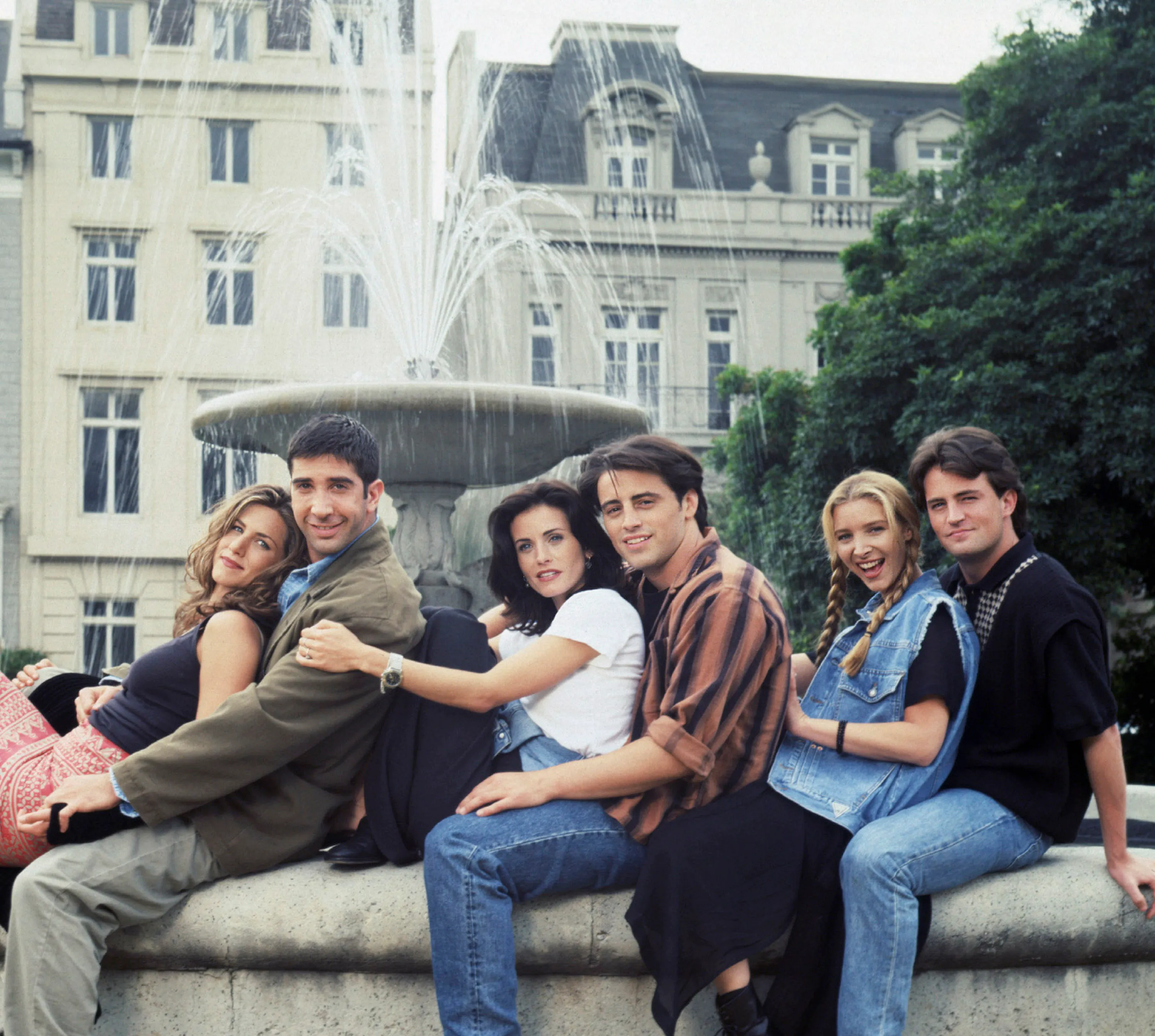 The cast of "Friends" sitting on the edge of a fountain, smiling at the camera.