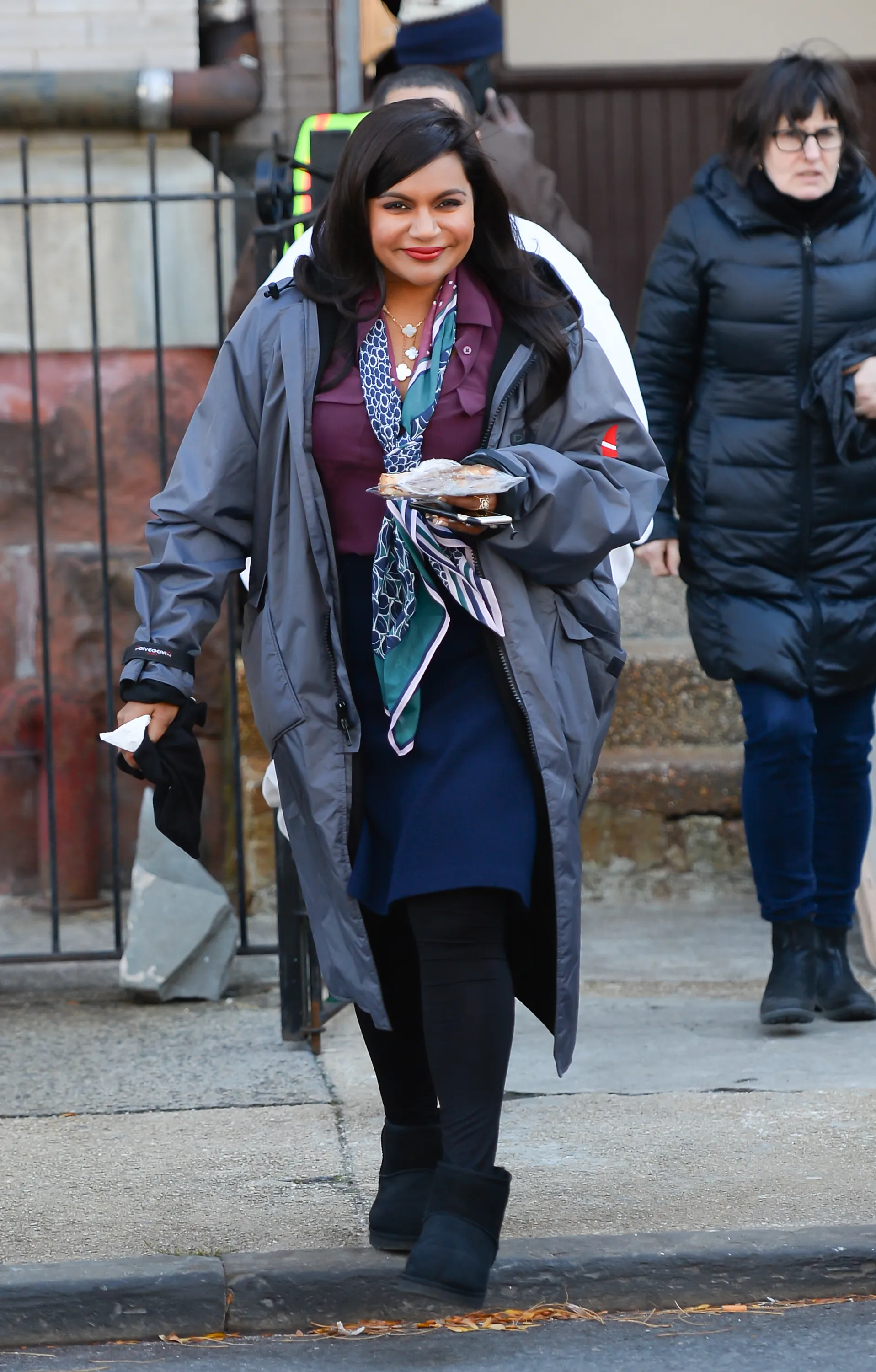 Mindy Kaling walking on the set of Ocean's Eight wearing a long coat, dark purple shirt, a scarf, and dark leggings.