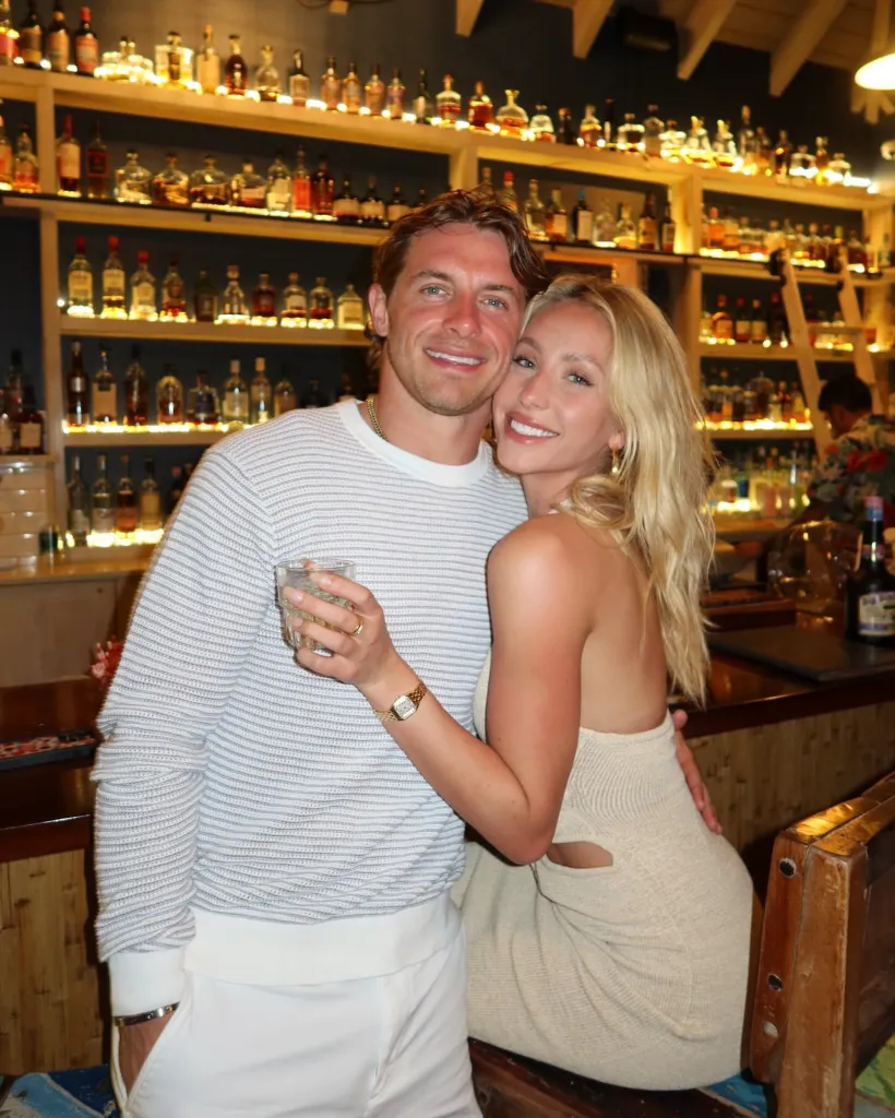 Alix Earle and Braxton Berrios smiling, Alix resting her arm around Braxton's waist in front of a bar.