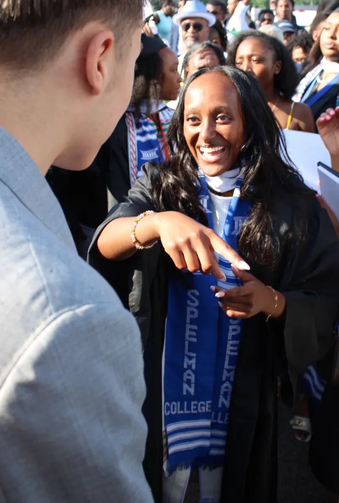 Zahara Jolie-Pitt smiles, wearing a graduation gown and blue Spelman College stole, pointing to someone off-camera.