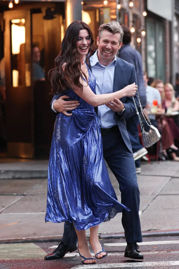Anne Hathaway in a shimmery blue dress embracing Patrick Brammall, both laughing, on the set of "The Devil Wears Prada II".