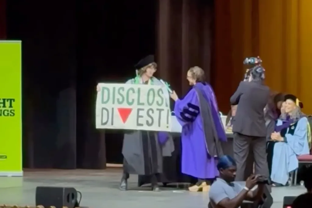 A graduating student holding a sign that reads "DISCLOSE DIVEST!" with a red inverted triangle, during a CUNY Law School graduation ceremony.