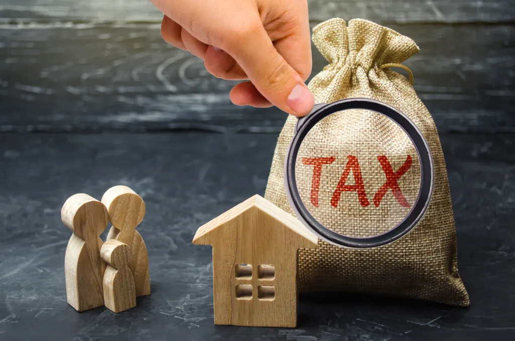 A hand holding a magnifying glass over a money bag labeled "TAX" with a wooden house and family figures nearby, illustrating property tax or real estate tax.