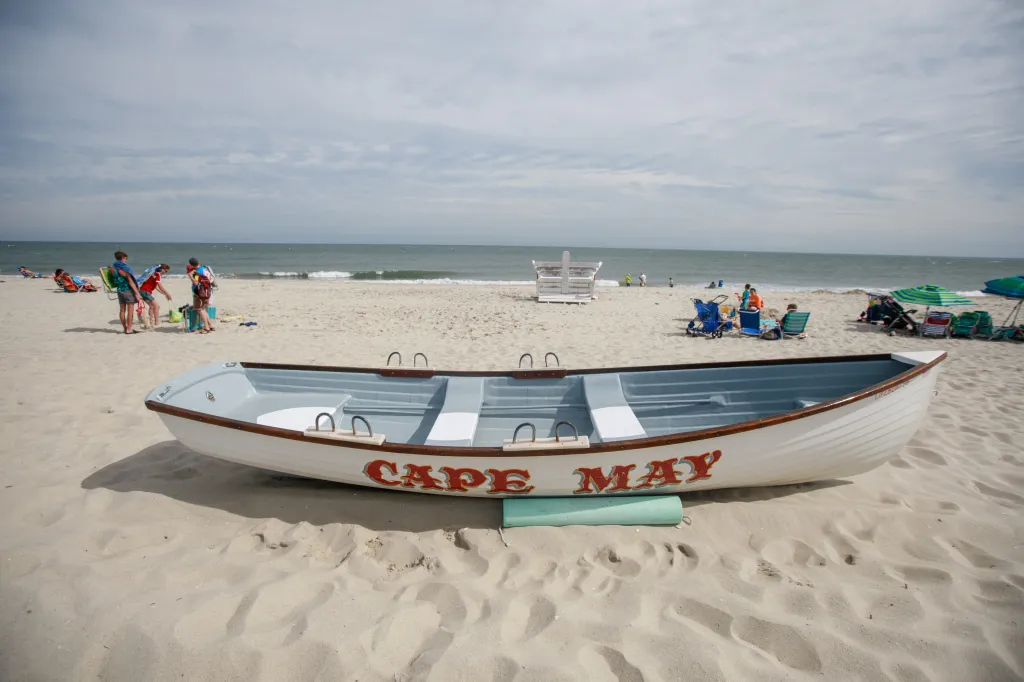 A white rowboat with "CAPE MAY" written on its side rests on the sandy beach of Cape May, New Jersey, with people and the ocean in the background.