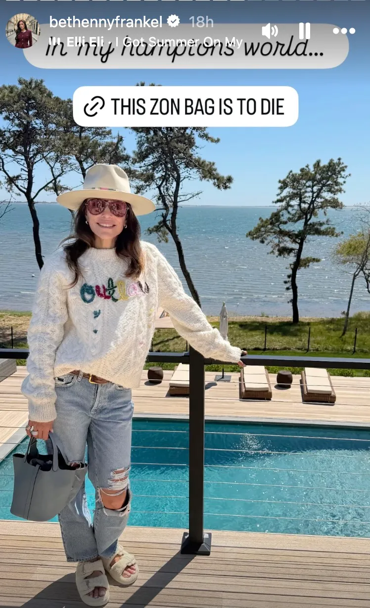 Bethenny Frankel posing in a cream sweater, ripped jeans, and a light gray handbag next to a pool overlooking the ocean.