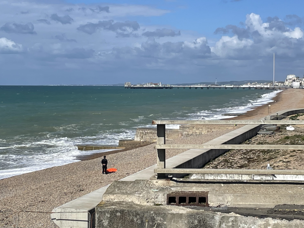 A bodybag on the Brighton beach, with a person standing nearby, and the sea in the background.