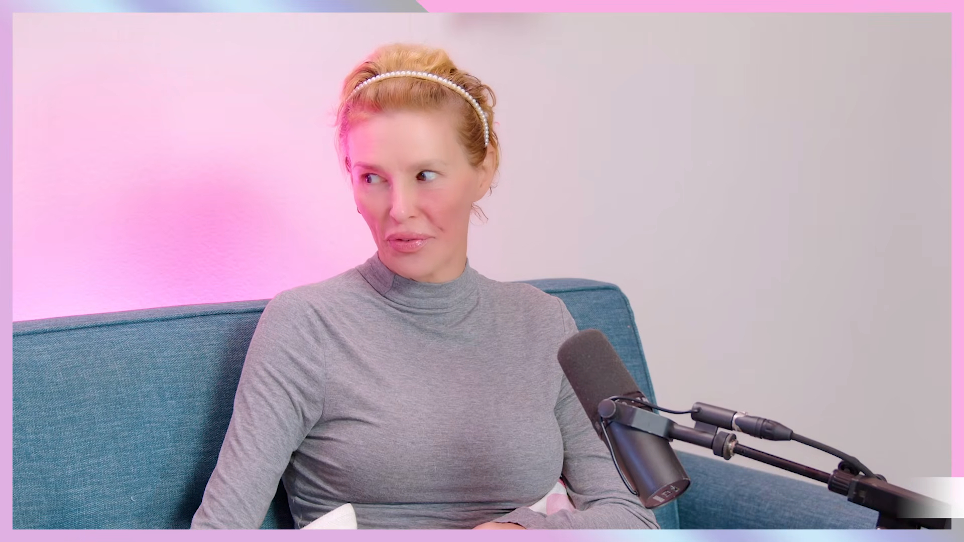 Brandi Glanville wearing a pearl headband and gray turtleneck, sitting on a blue couch next to a microphone.