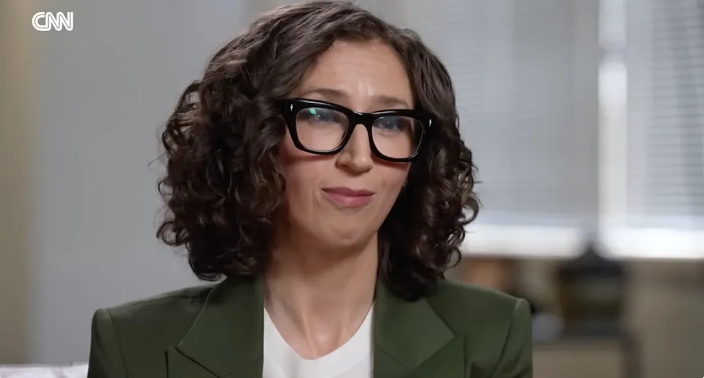 Brooke Nevils, wearing glasses and a green blazer, looks thoughtfully at the camera.