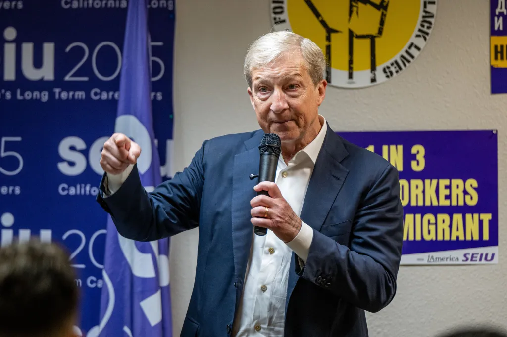 Tom Steyer speaking into a microphone, gesturing with his right hand, with a SEIU 2015 flag and a "BLACK LIVES" poster in the background.