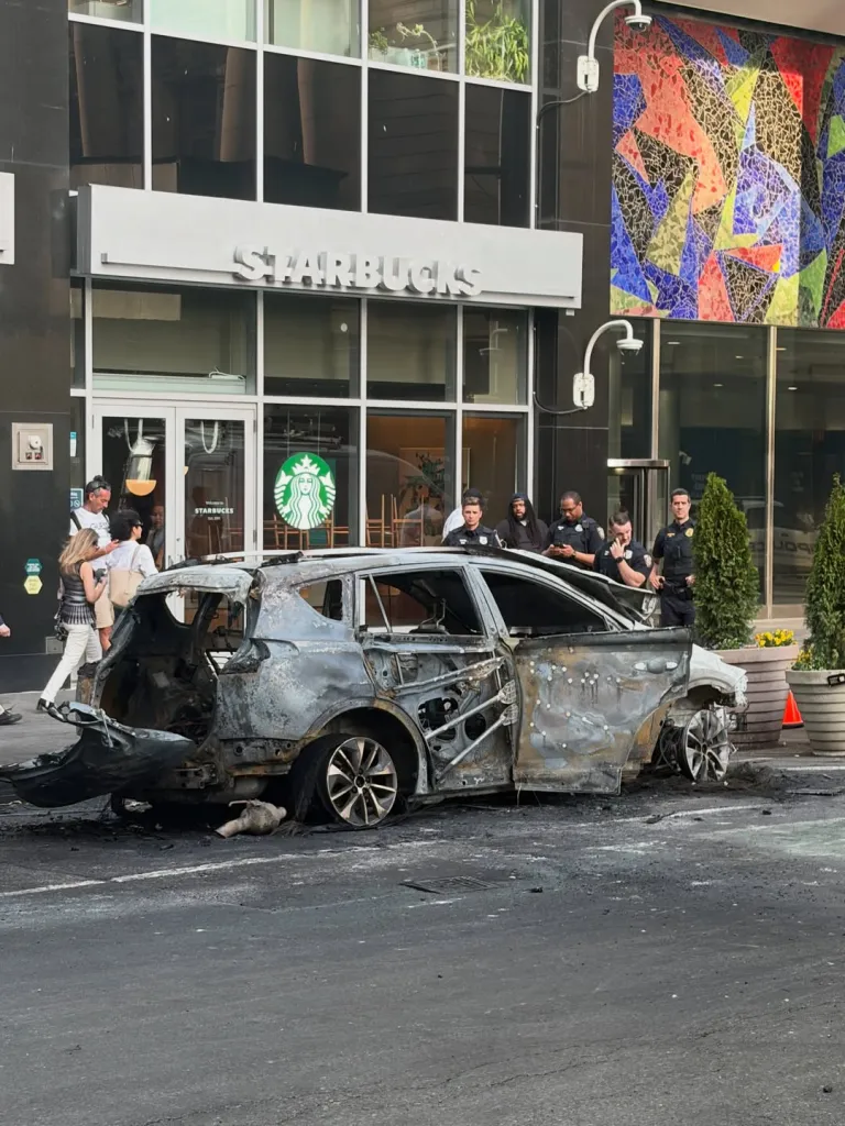 A burned-out car on a city street in front of a Starbucks, with people and police officers nearby.