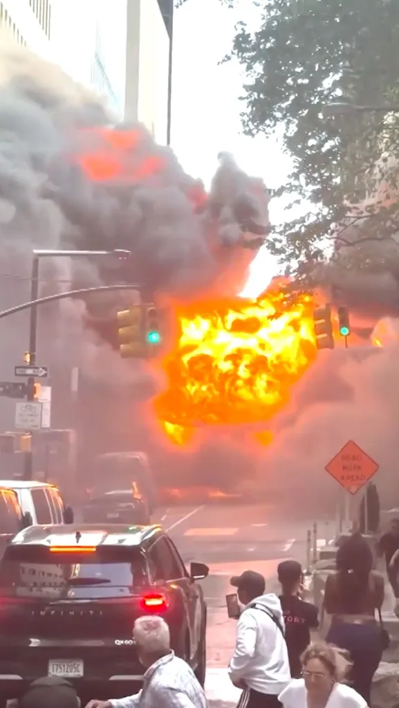 A car engulfed in a large fireball and thick black smoke on a city street, with onlookers in the foreground.
