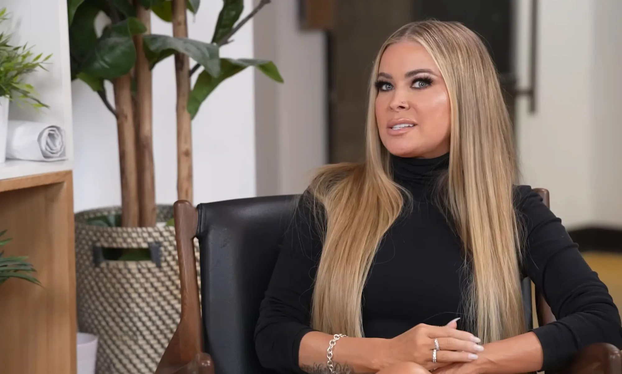 Carmen Electra sitting in a black chair with long blonde hair, looking towards the right.