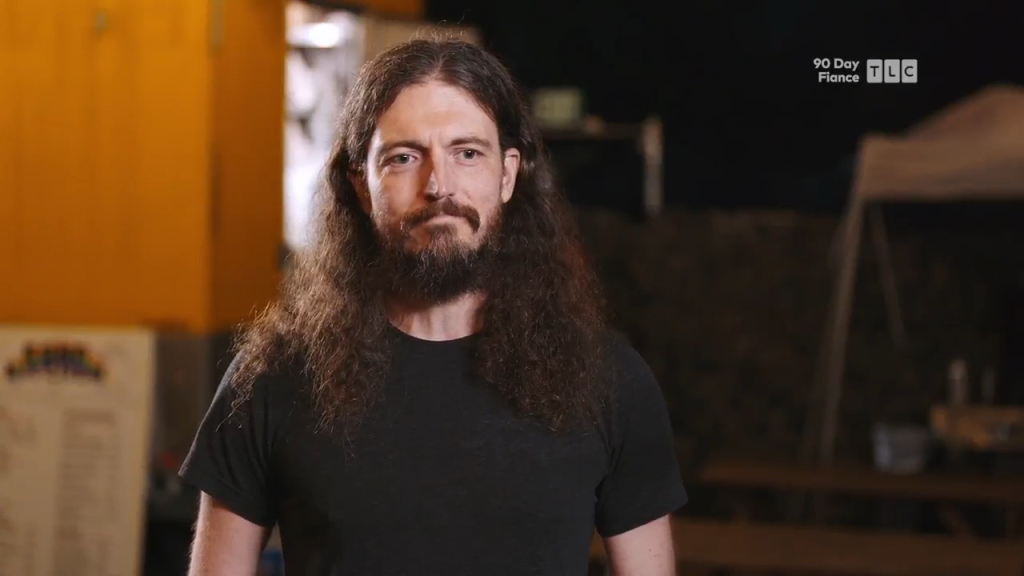 A man with long curly brown hair and a beard looks seriously at the camera.