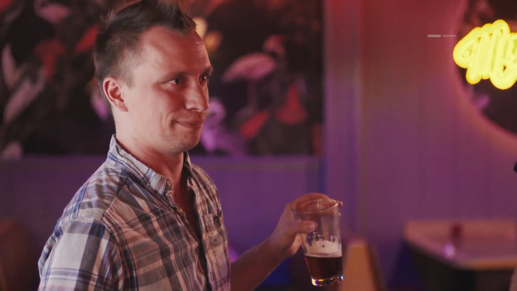 Josh holding a glass of beer, looking slightly to the left, with neon lights in the background.