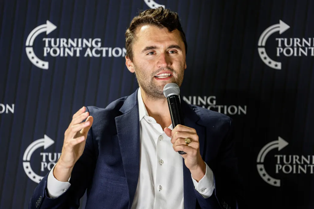 Charlie Kirk speaking into a microphone, with a Turning Point Action logo in the background.
