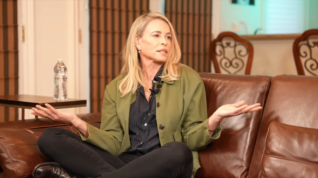 Chelsea Handler sitting on a brown leather couch, speaking with her hands open.